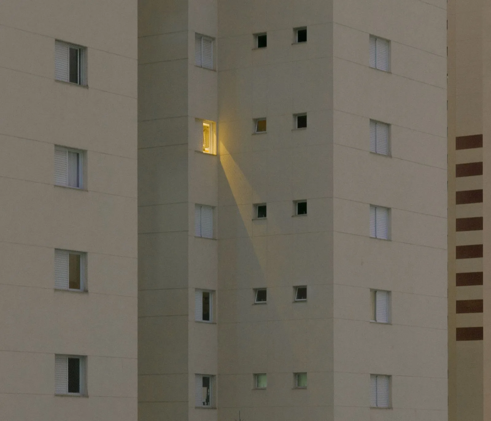 One lit window on the pale wall of a tall apartment building