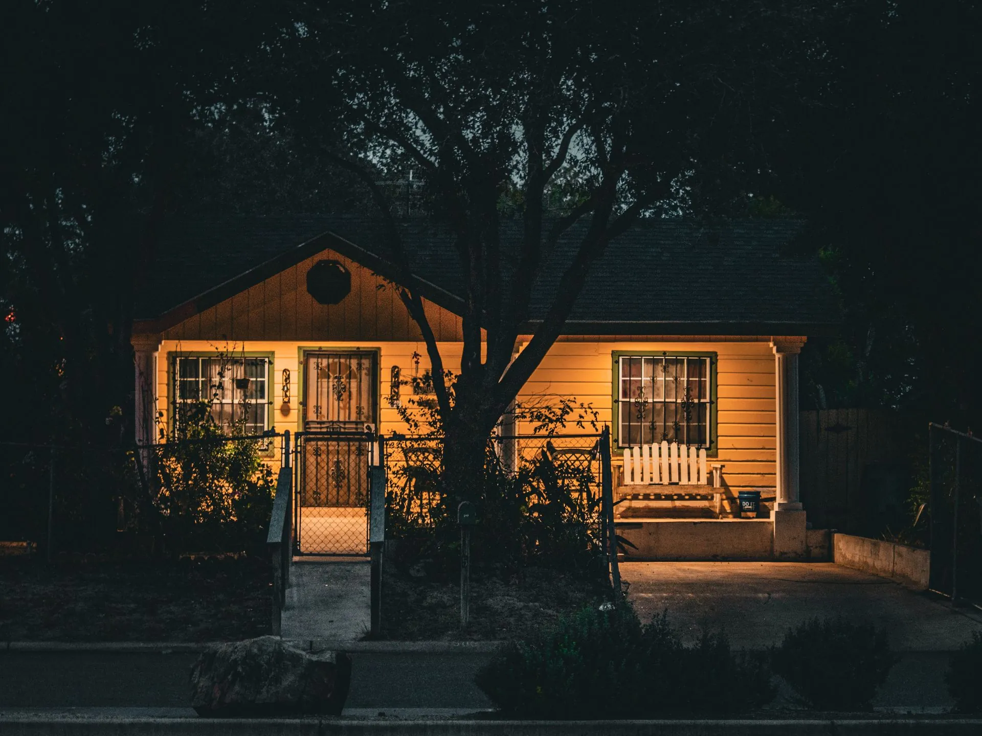 A small house at night with warm light in every window and a lamp glowing over the porch
